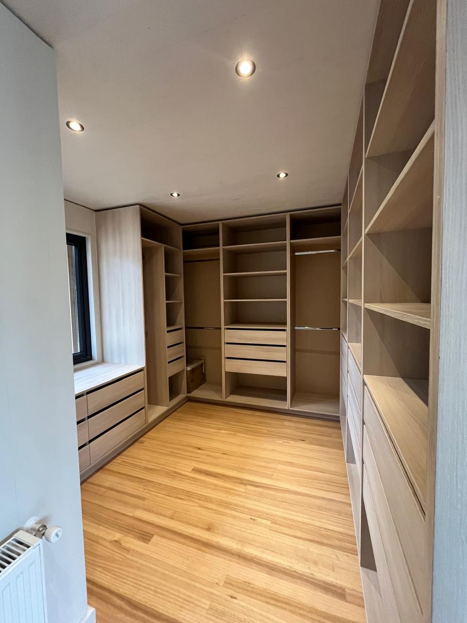 Walk-in closet a medida en L, con cajoneras, repisas abiertas, barras de colgar y banca bajo la ventana