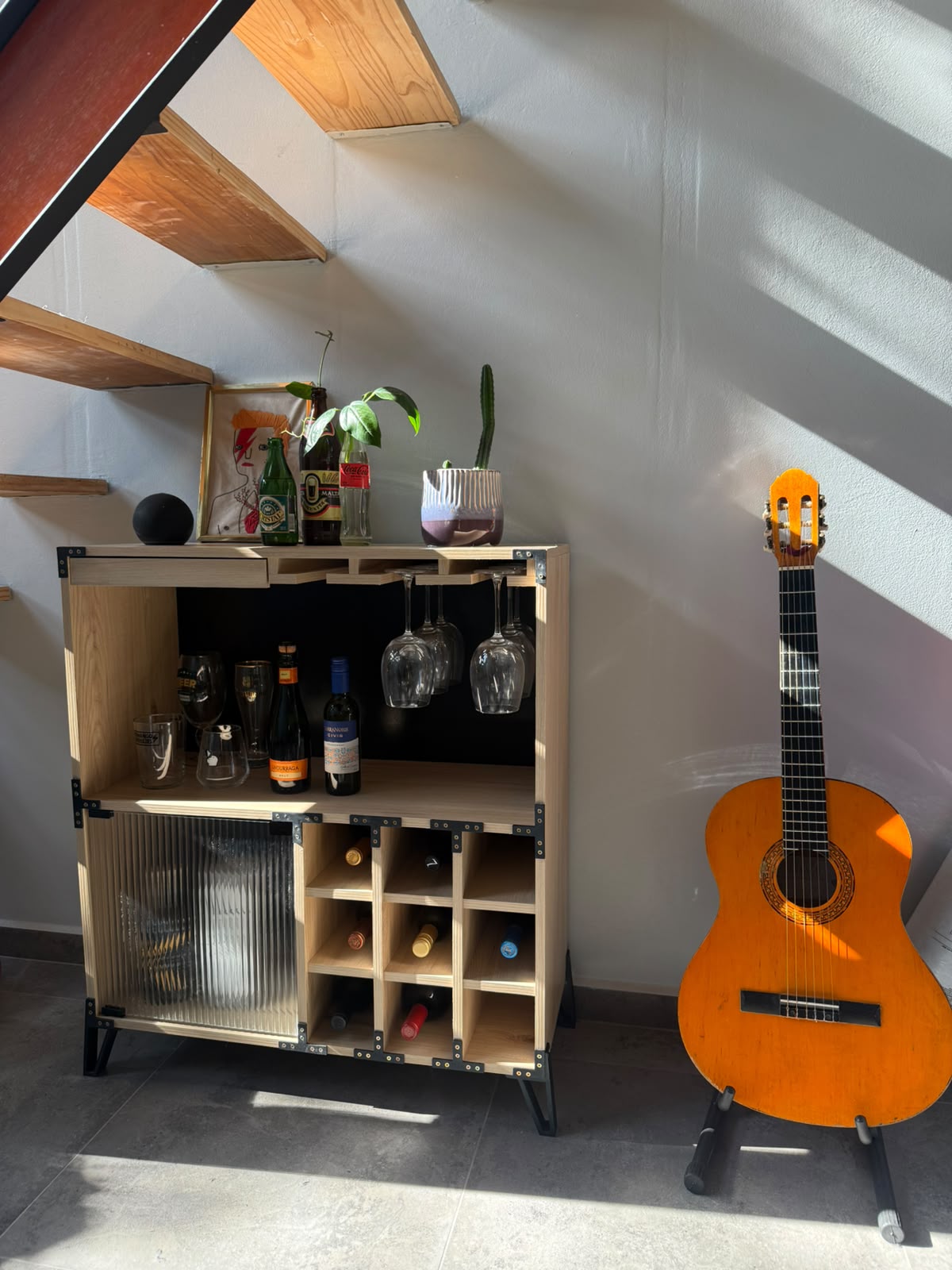 Mueble bar Bar Rock de El Taller Once instalado bajo una escalera, con botellas, copas colgando del copero y botellero cargado